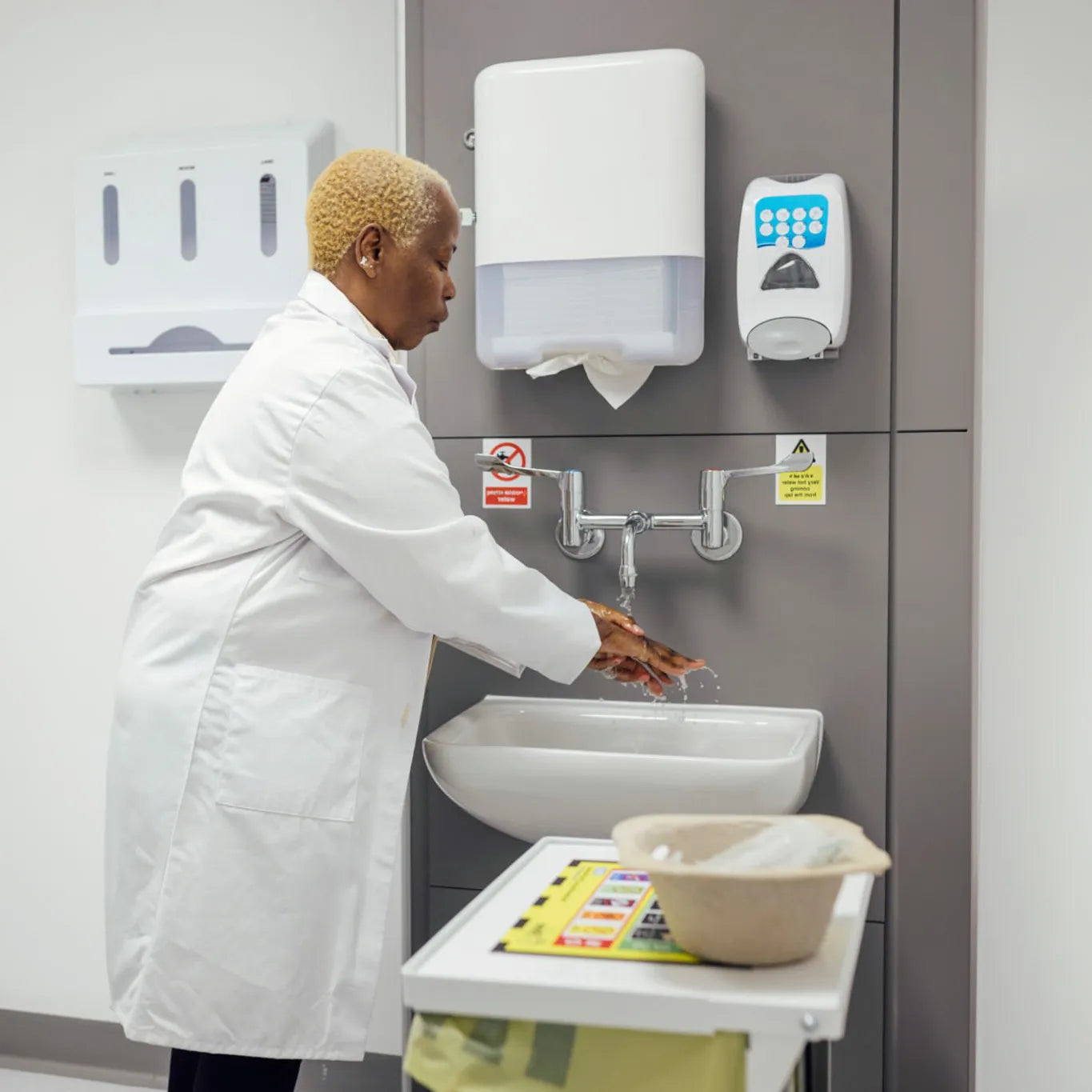 Close-up of a person washing hands under a manual faucet in a standard healthcare sink, illustrating the contamination risks of traditional sink designs in clinical settings.