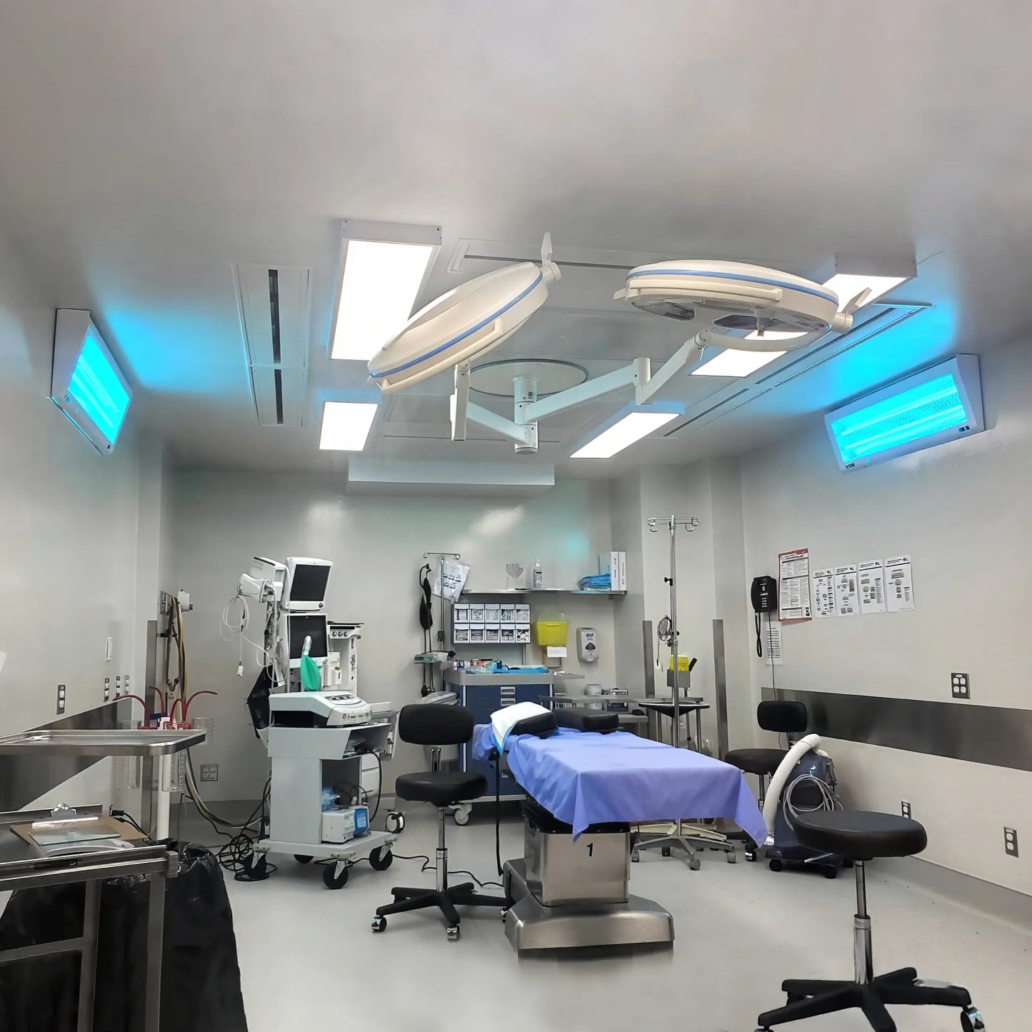 ASEPT 1X MAX sanitizers mounted on opposite walls of a hospital operating room, glowing blue during an active disinfection cycle.