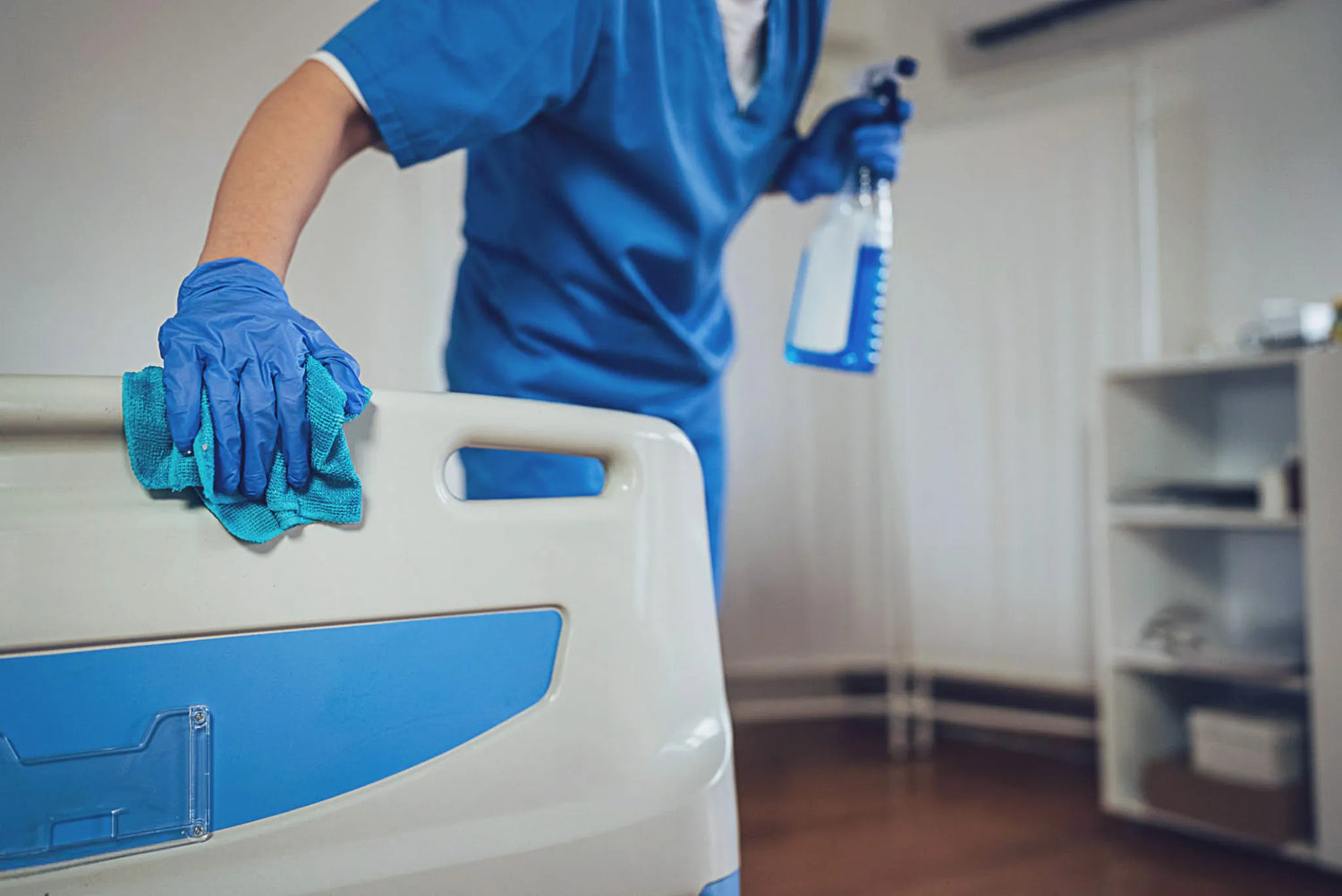 A healthcare worker manually disinfecting a hospital bed with gloves and spray, illustrating the labour-intensive and error-prone nature of traditional cleaning methods.